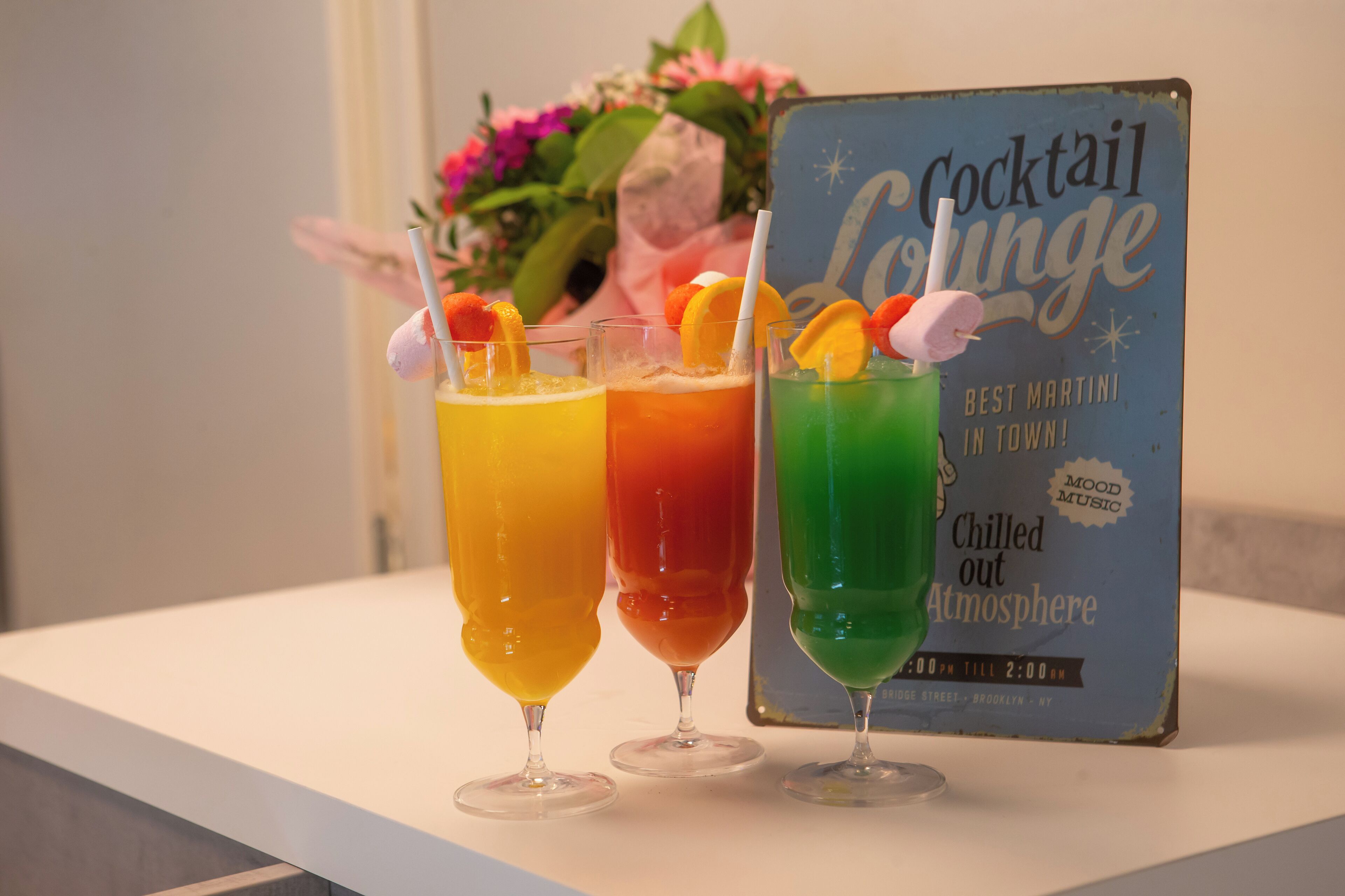 Three cocktails with fruit garnishes on a table, a sign for a cocktail lounge, and a bouquet of flowers in the background.