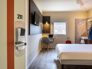 A hotel room with a bed, a desk with a laptop, a chair, a TV mounted on the wall, a window with a view, and a door with a number. - ibis budget Haguenau (Haguenau)