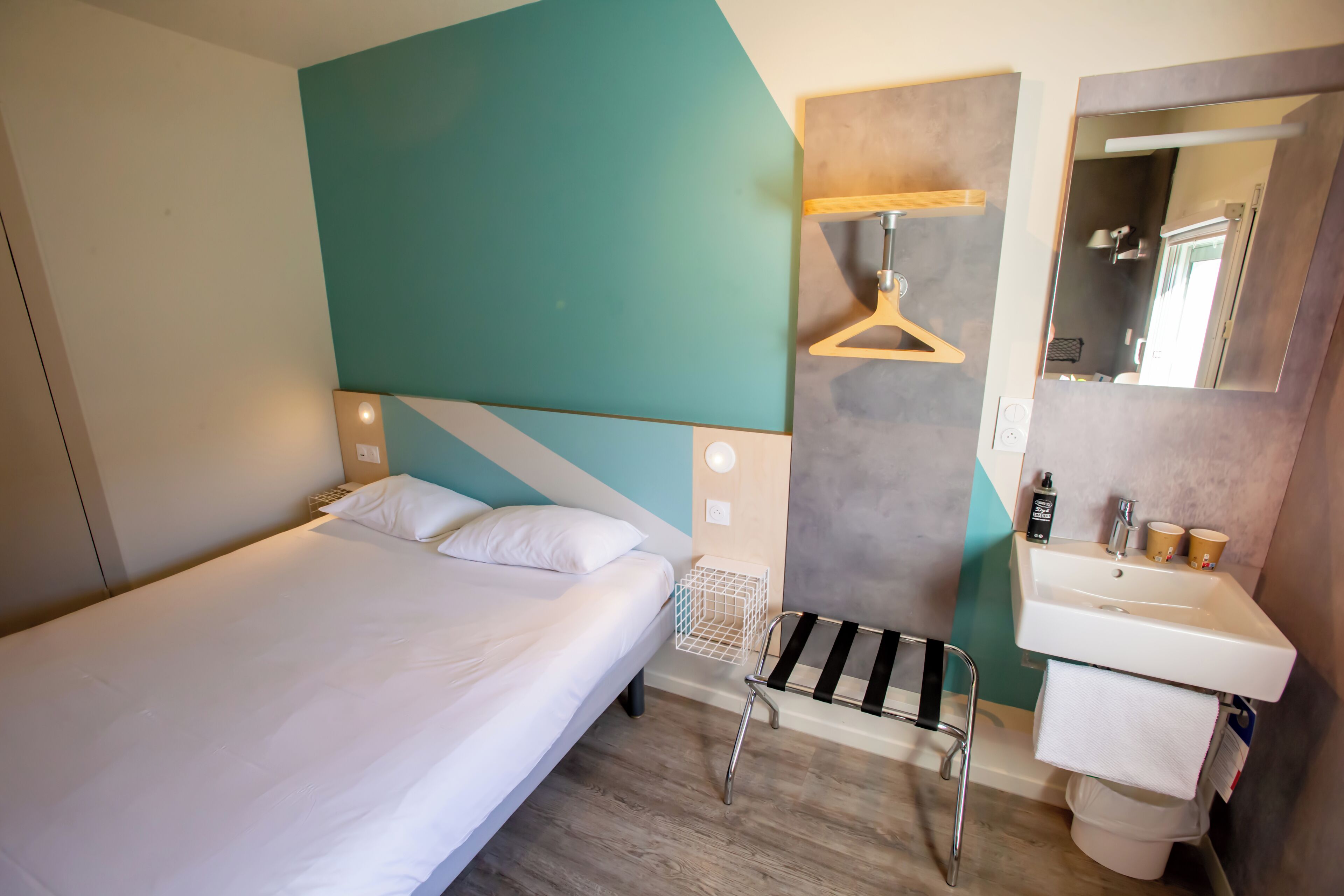 Habitación de hotel con una cama individual, una mesita de noche, una lámpara colgante, un baño con lavabo y espejo, y un perchero para toallas.