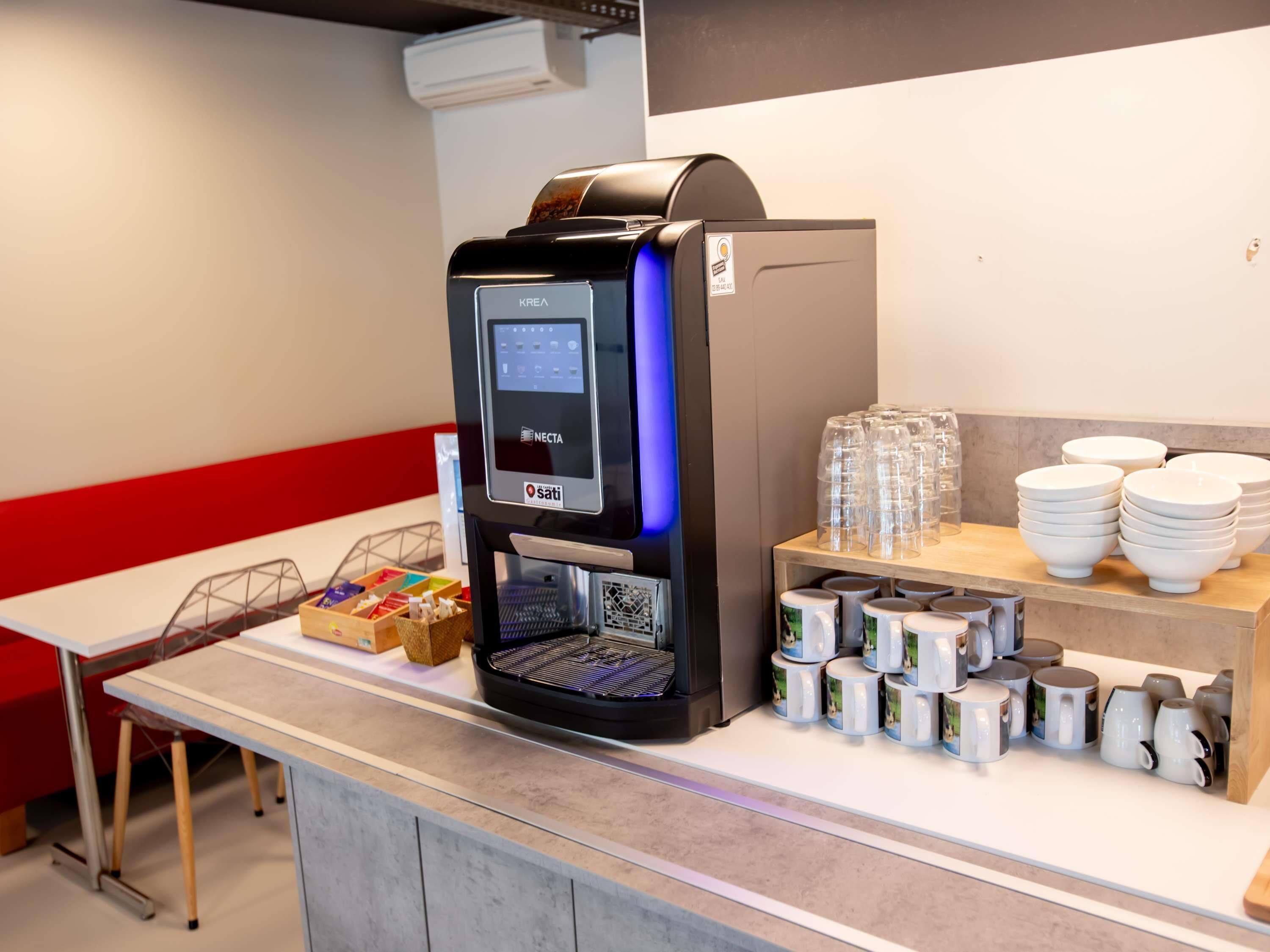 Máquina de café com visor digital, espaço com xícaras e pires, e pequena seleção de lanches.