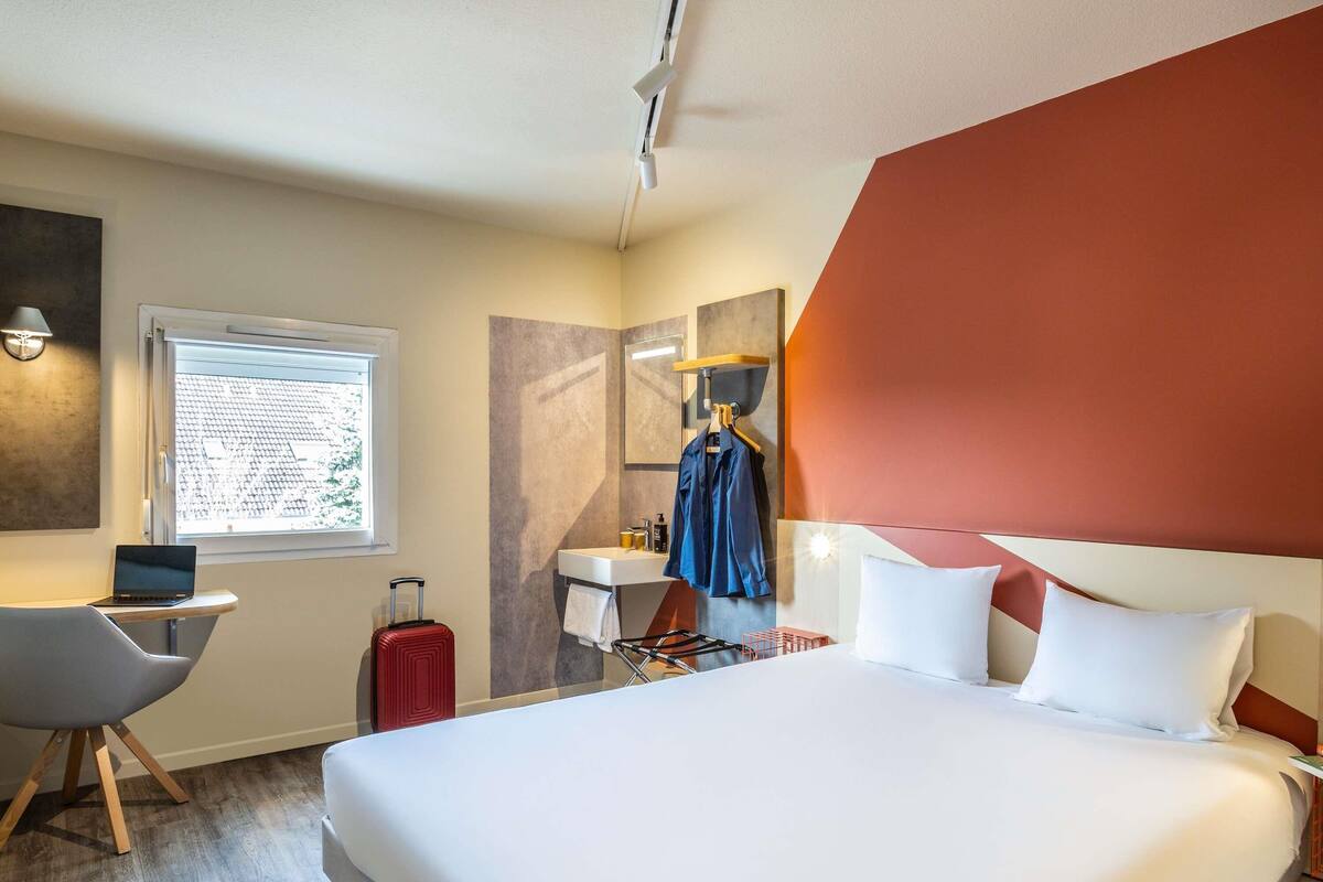 Een hotelkamer met een bed, een bureau met een laptop, een koffer, een raam met uitzicht, een badkamer met een spiegel en een kapstok.
