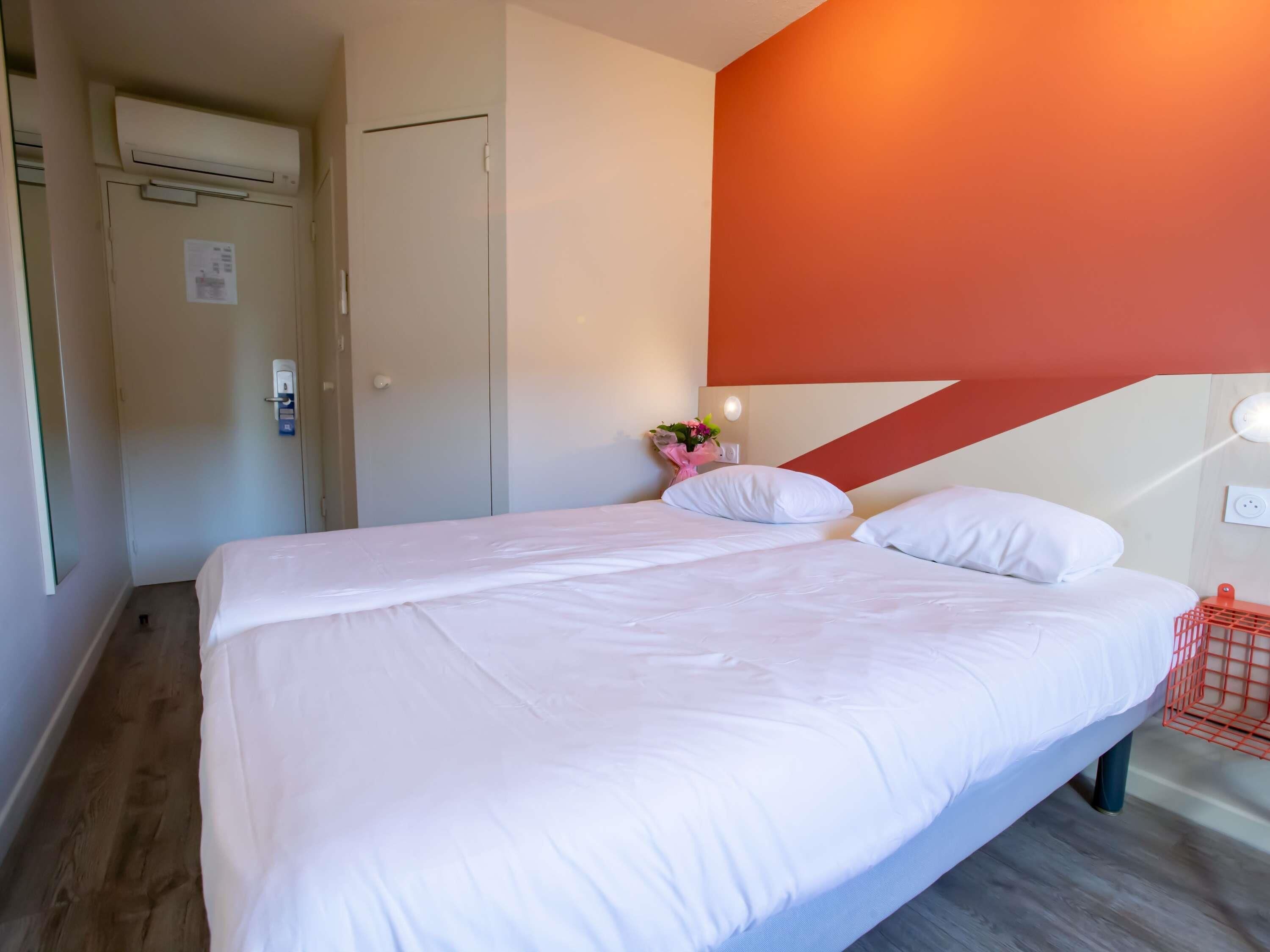 Un lit double avec du linge de lit blanc, un plancher en bois, une porte avec un interrupteur et un mur agrémenté d’une touche de couleur rouge.
