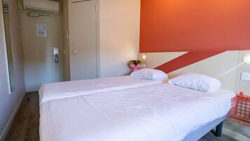 Un lit double avec du linge de lit blanc, un plancher en bois, une porte avec un interrupteur et un mur agrémenté d’une touche de couleur rouge.