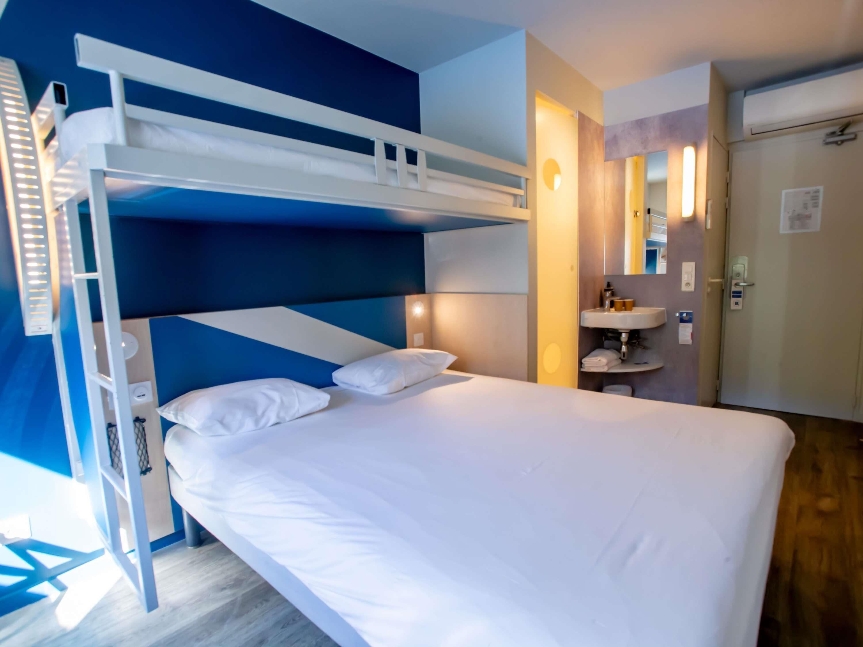 Una cama individual con ropa de cama blanca, una mesita de noche pequeña y un espejo sobre el lavabo en una habitación con paredes azules y beige.