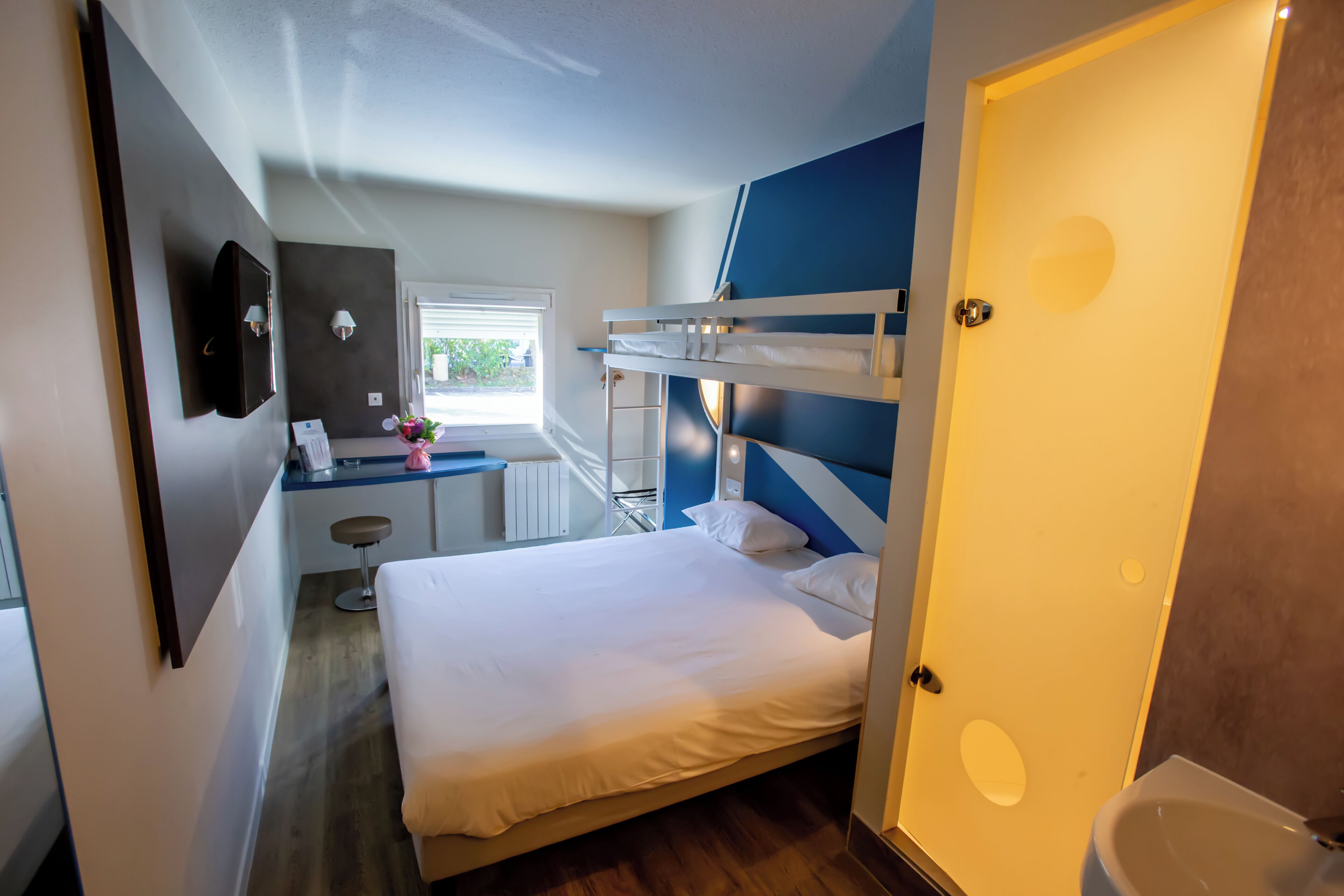 A bedroom with a bunk bed, a desk with a chair, a small stool, and a window.