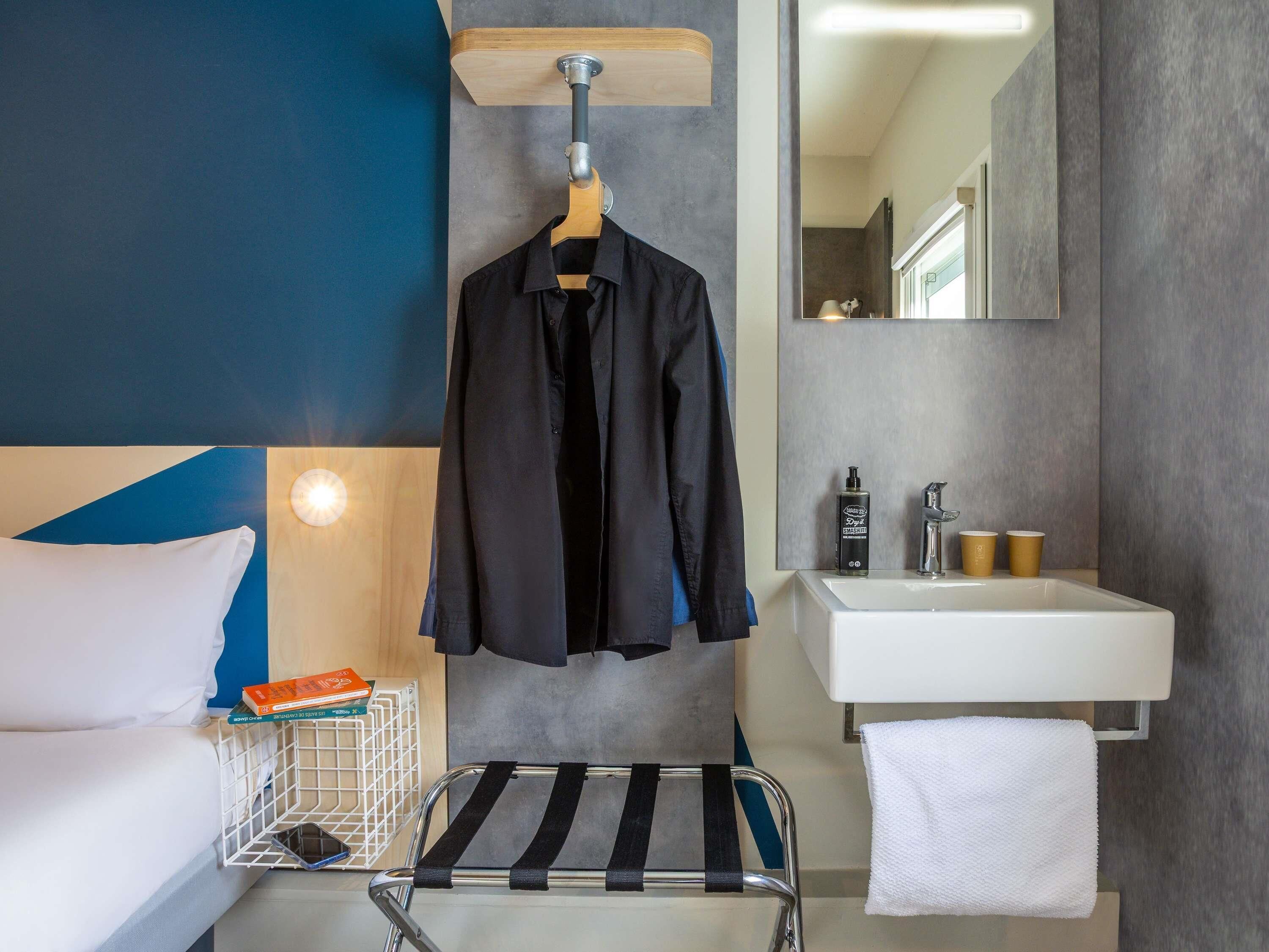 A hotel room with a bed, a hanging coat rack, a bedside table with a book and a phone, a towel, a small trolley, and a bathroom with a sink and mirror.