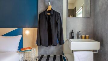 A hotel room with a bed, a hanging coat rack, a bedside table with a book and a phone, a towel, a small trolley, and a bathroom with a sink and mirror.