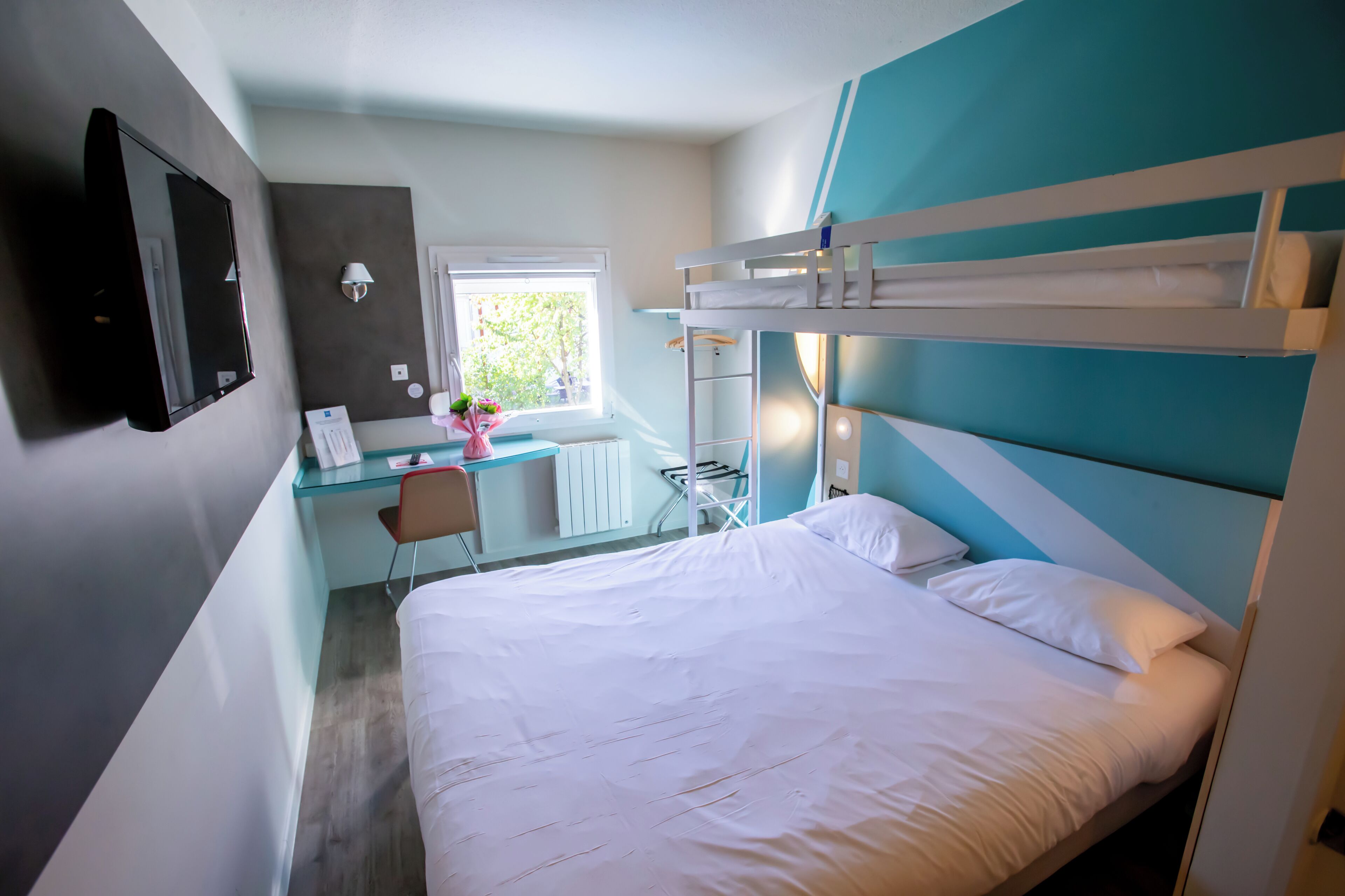 A bedroom with a bunk bed, a desk with a chair, a window, and a wall-mounted TV.