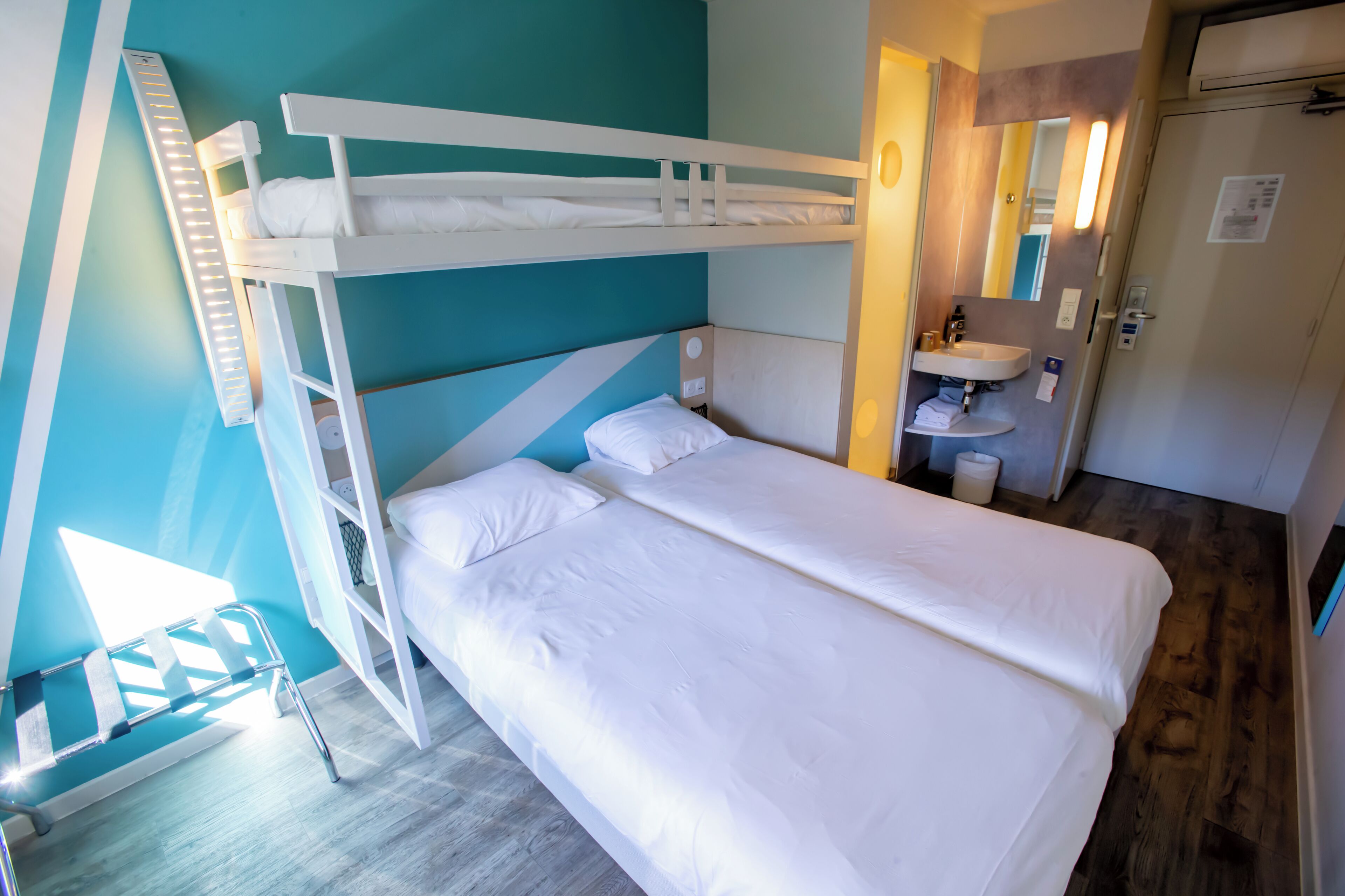 A bunk bed with white bedding, a bedside table, and a mirror. There is a bathroom with a sink and a toilet visible in the background.