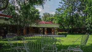 A covered outdoor seating area with metal chairs and a table, surrounded by a well-maintained lawn and trees. - Matilinda Lodge (Bulawayo)