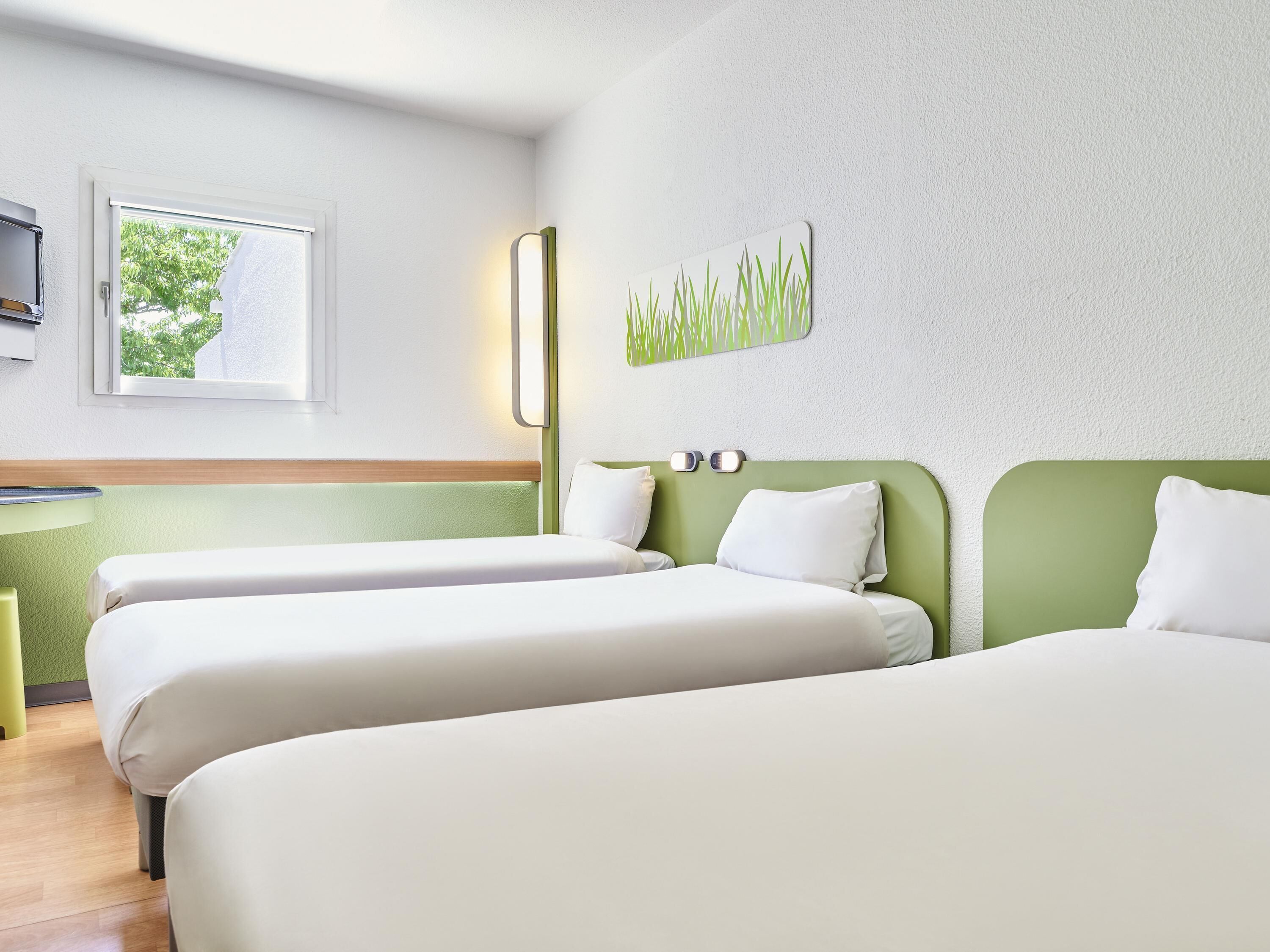 A hotel room with two beds, a small desk, a window with a view of trees, and a wall-mounted artwork.