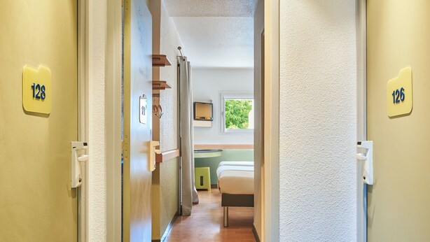 A hallway with doors labeled 128 and 126, leading to a room with a bed, a window, and a small stool. - ibis budget Versailles Coignières (Coignieres)