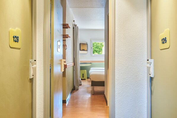 Un pasillo con puertas identificadas con los números 128 y 126, que conduce a una habitación con una cama, una ventana y un pequeño taburete.