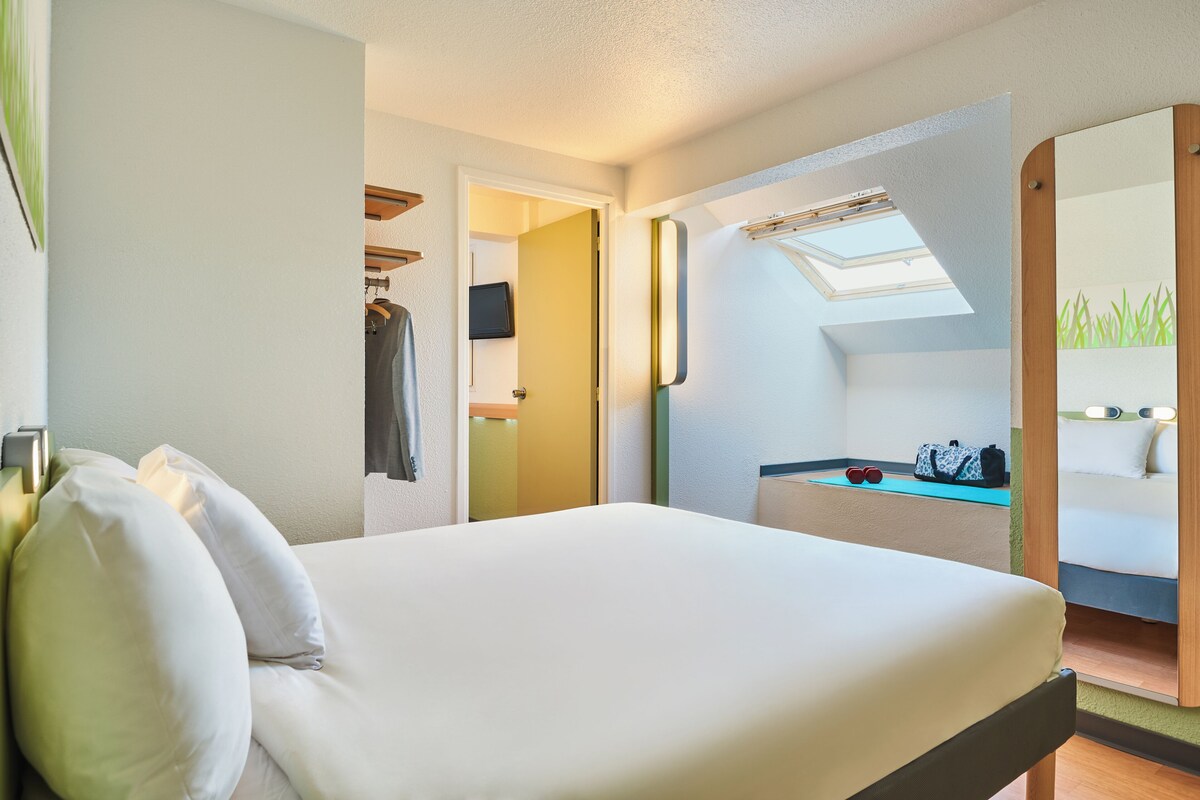 A hotel room with a bed, a mirror, a desk, and a skylight.