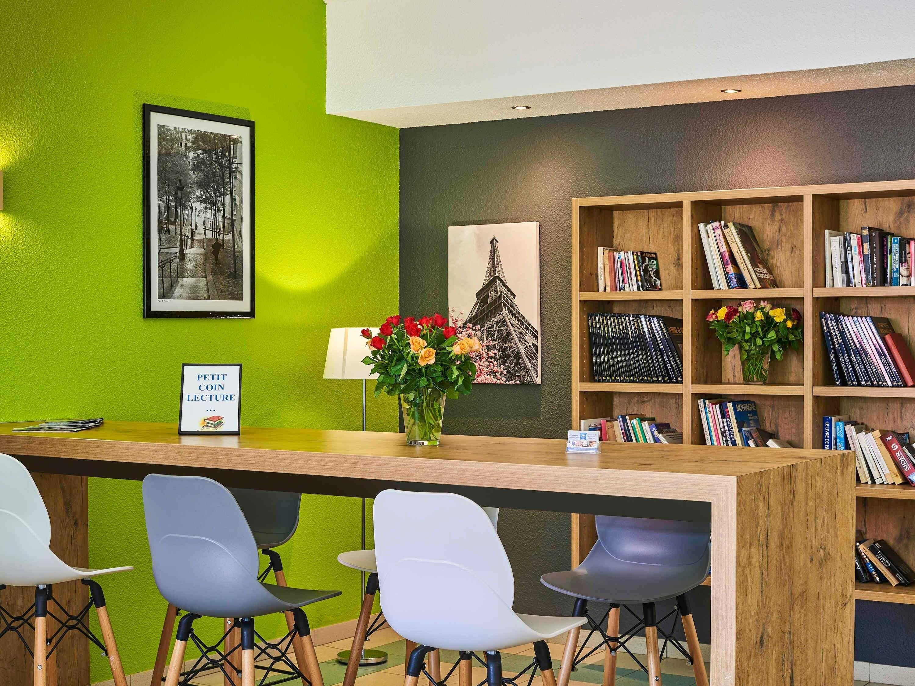 Un mostrador de recepción con una pared verde, un cuadro con la Torre Eiffel, una estantería con libros y un jarrón con flores.