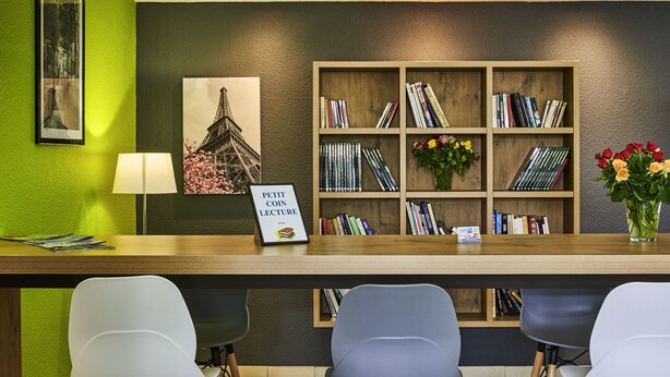 A reception desk with a wooden bookshelf, a framed picture of the Eiffel Tower, a sign reading "PETIT COIN LECTURE," and a vase with flowers.