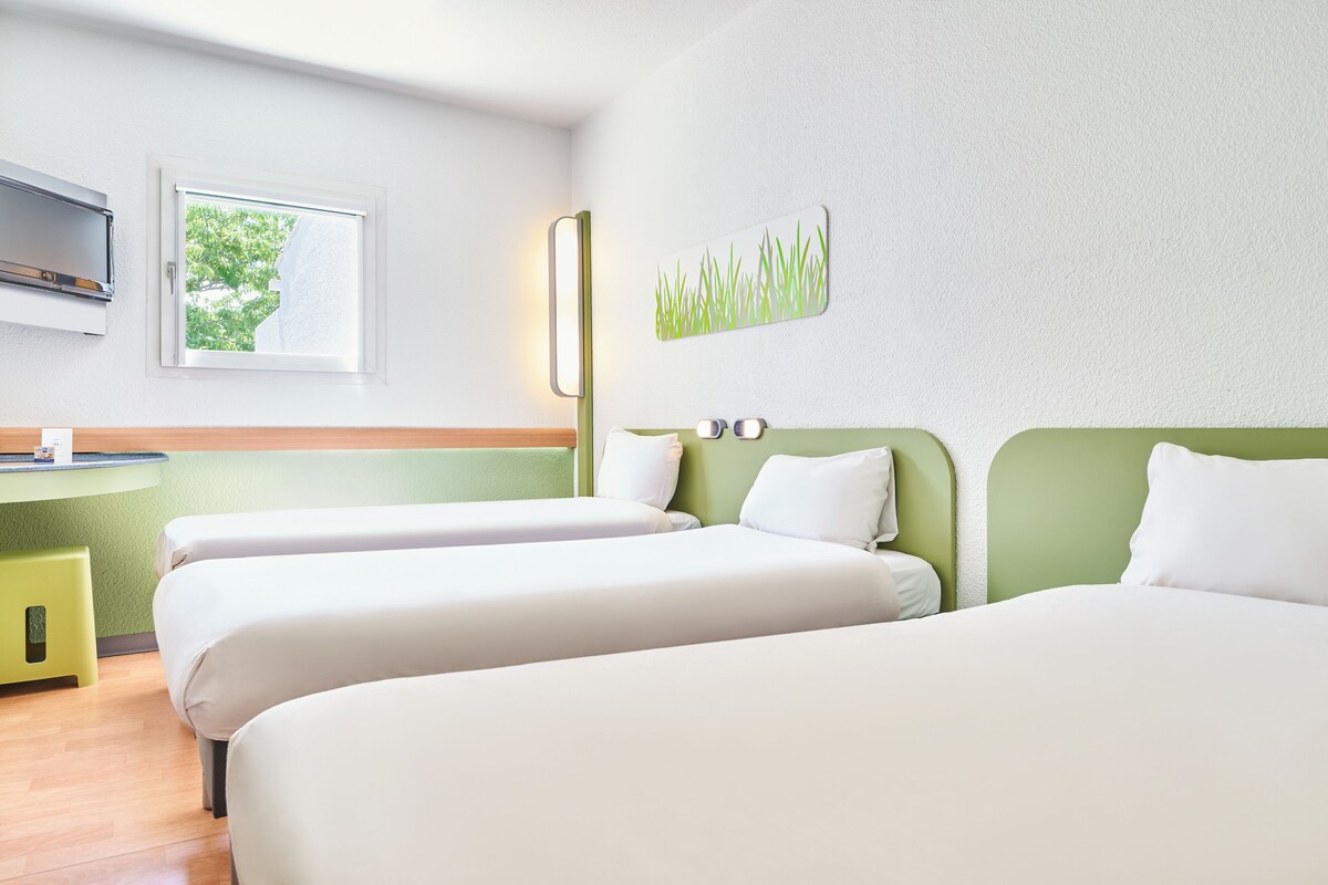 A hotel room with two beds, a small desk, a window with a view of trees, and a wall-mounted artwork.