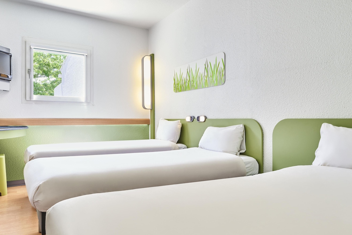 A hotel room with two beds, a small desk, a window with a view of trees, and a wall-mounted artwork.