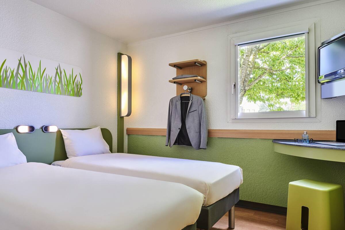 A hotel room with two beds, a desk with a computer, a TV, and a window with a view of trees.