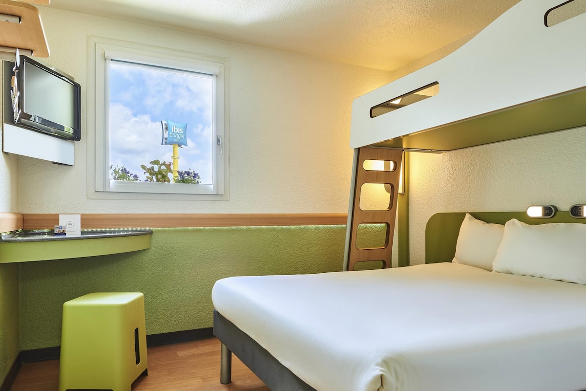A compact hotel room with a bed, a desk, a small stool, a window with a view, and a television.