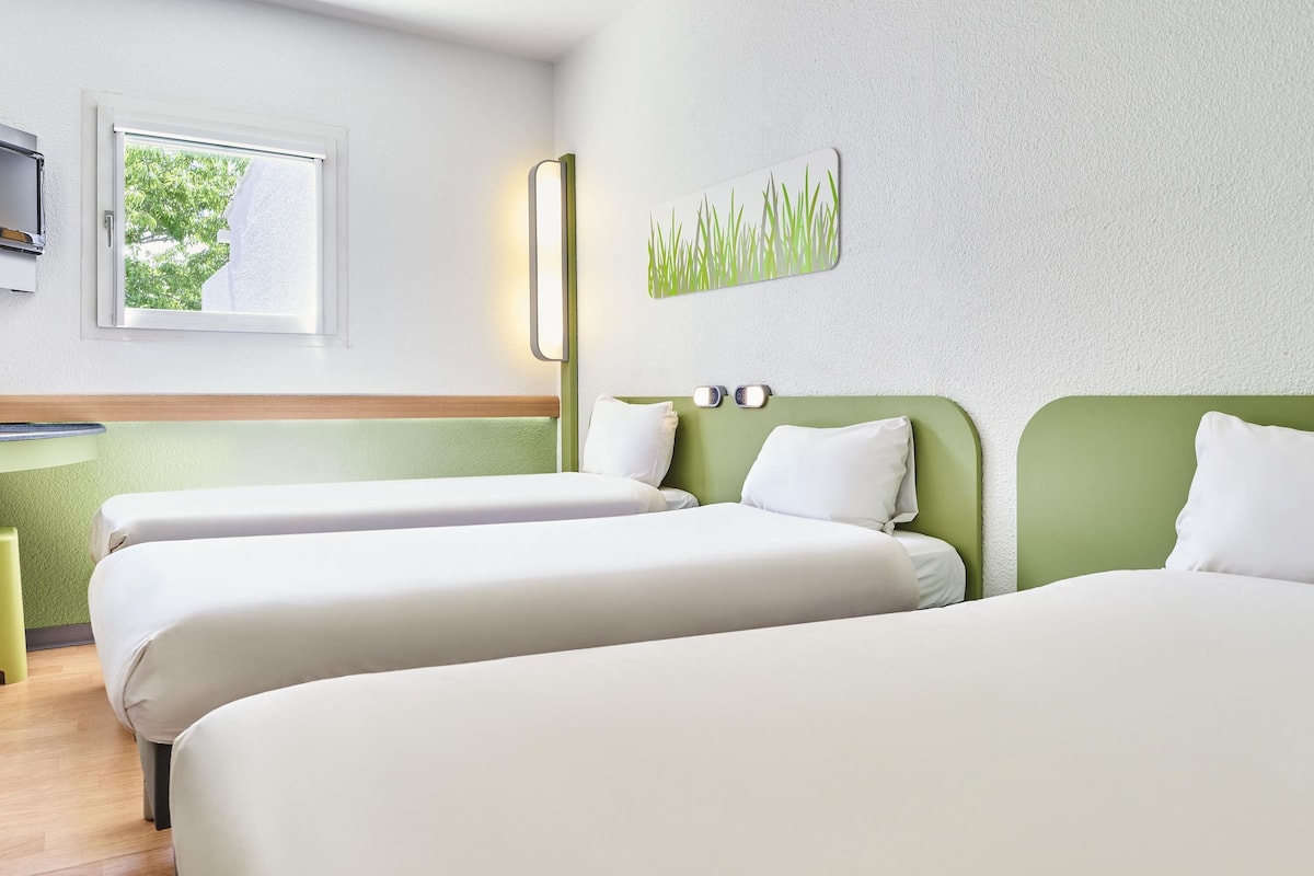 A hotel room with two beds, a small desk, a window with a view of trees, and a wall-mounted artwork.