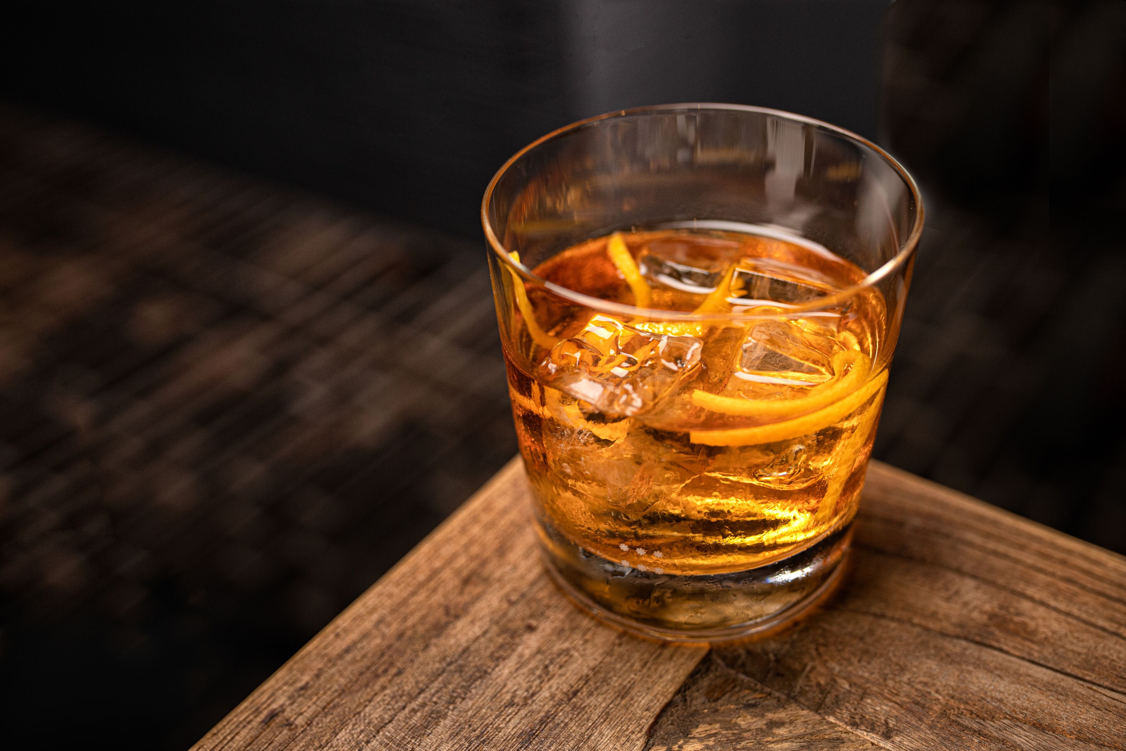 A glass of amber liquid with ice and a slice of orange peel, placed on a wooden surface.