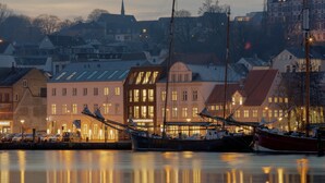 A harbor with boats docked, buildings with traditional architecture, and a calm water surface reflecting the structures. - Hotel Hafen Flensburg (Flensburg)