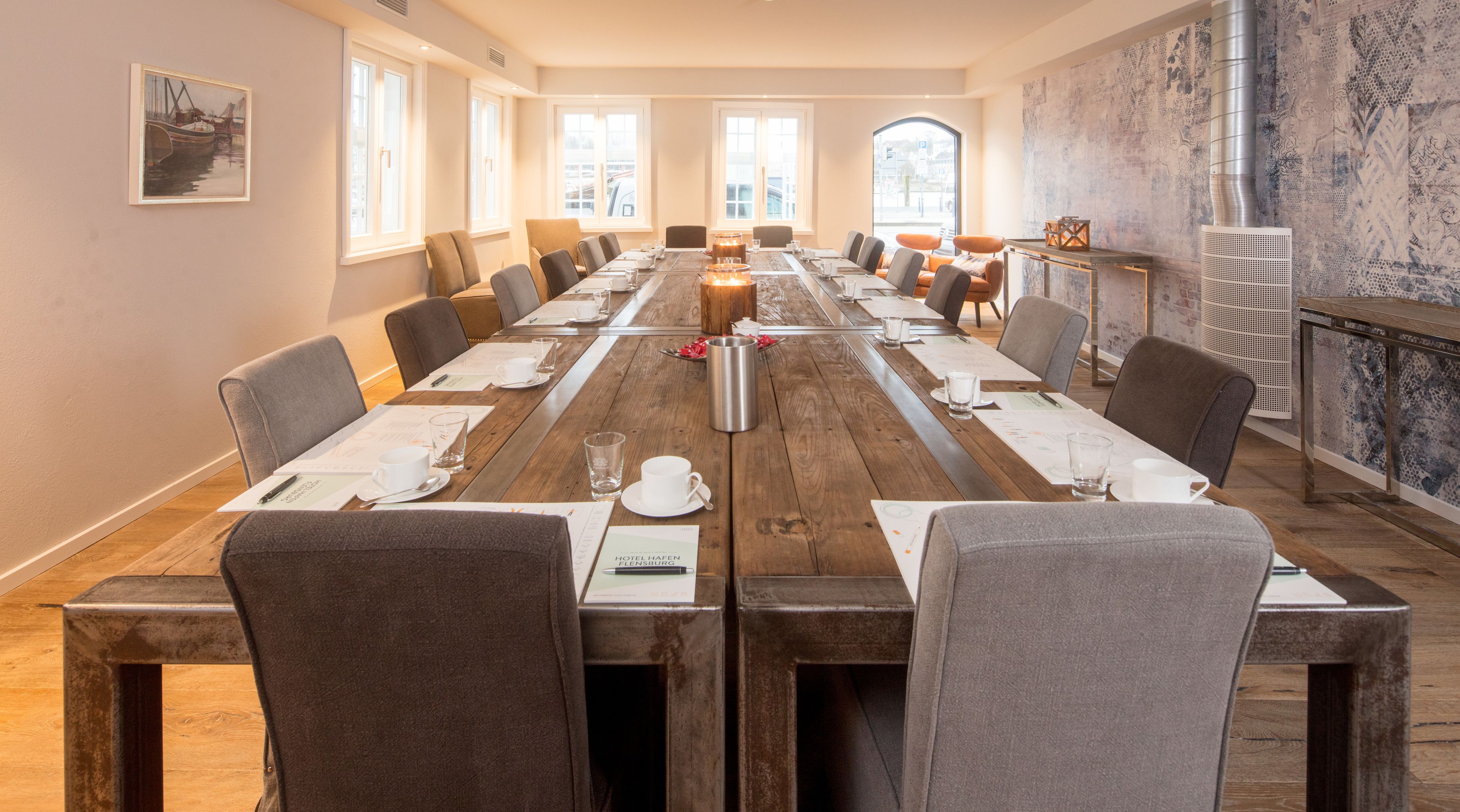 A conference room with a long wooden table, grey upholstered chairs, and a large window with a view of buildings.