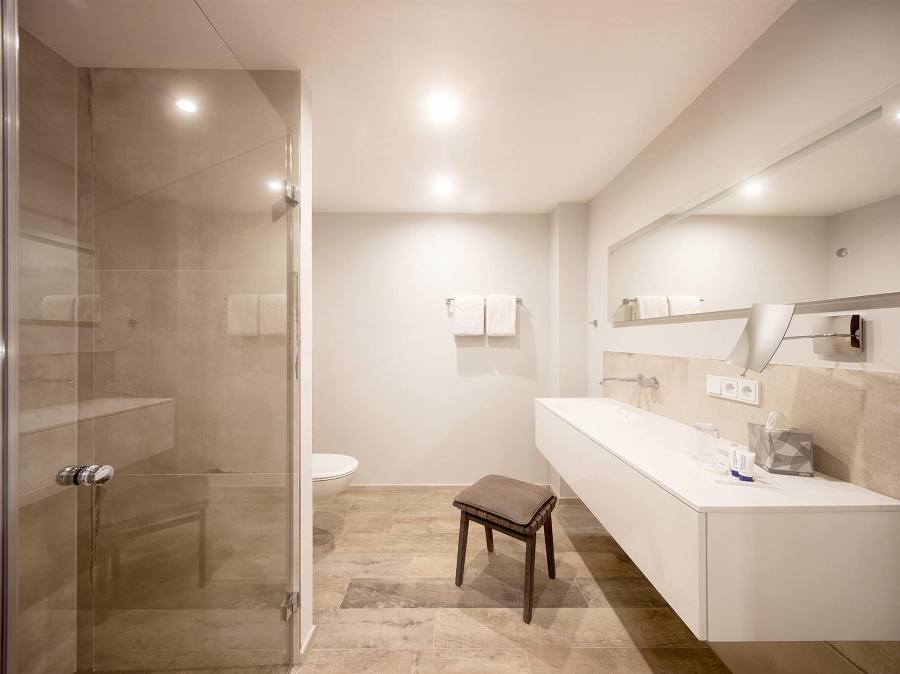 Et moderne badeværelse med et glas brusekabine, et hvidt toilet, et vaskebord med spejl og en lille skammel.
