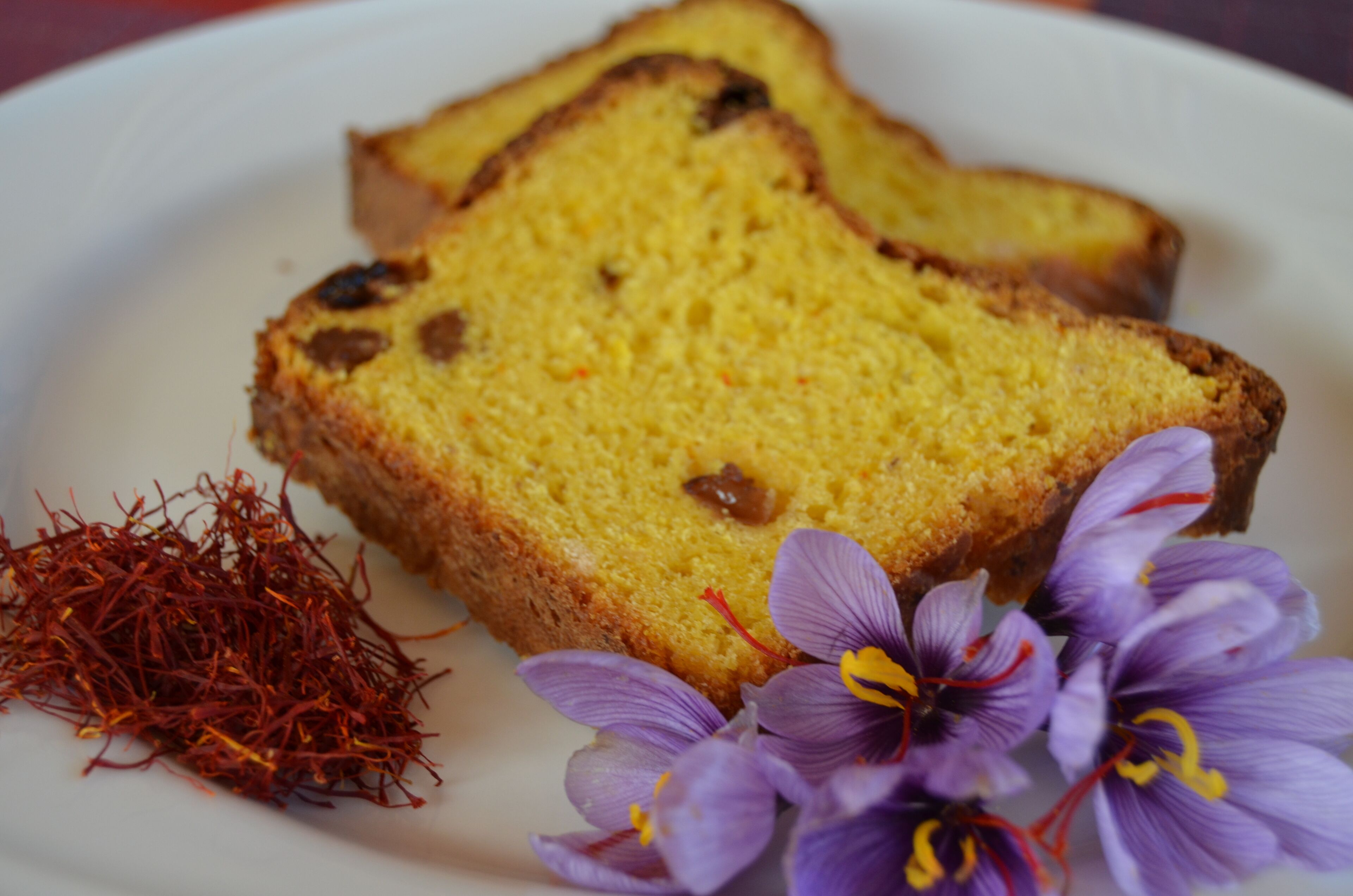 Gelbe Kuchenstücke mit Rosinen und Safranfäden auf einem Teller, begleitet von violetten Blüten.