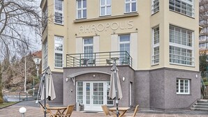 A multi-story building with the name "AKROPOLIS" on the facade. It features a ground-level entrance with a glass door, a balcony with outdoor seating, and a paved area with tables and chairs. - Apart Hotel Akropolis (Karlovy Vary)