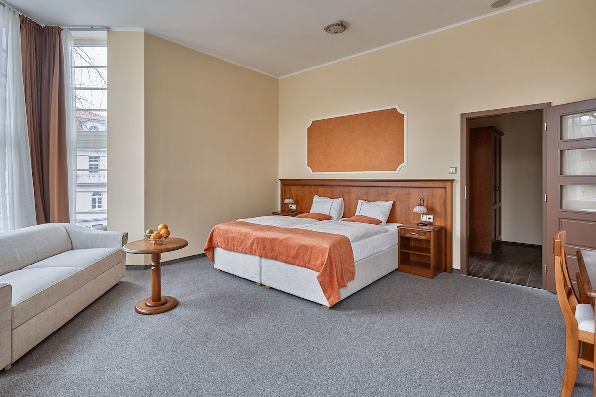 Quarto de hotel com uma cama, um sofá, uma mesinha redonda com frutas, um criado-mudo e uma cadeira de madeira.