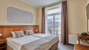 A hotel room with a large bed, a wooden headboard, a bedside table with a lamp, a desk with a telephone, and a view of the outdoors through a window. - Apart Hotel Akropolis (Karlovy Vary)