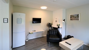 Un espacio habitable compacto con un refrigerador blanco, un televisor de pantalla plana montado en la pared, un sillón de cuero negro, una mesita de madera y una lámpara de pie.