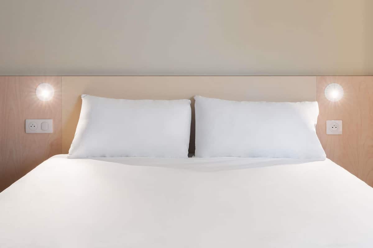 Una cama bien hecha con sábanas blancas, dos lámparas fijadas a la pared y un cabecero de madera.