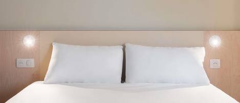 A neatly made bed with white linens, two wall-mounted light fixtures, and a wooden headboard.