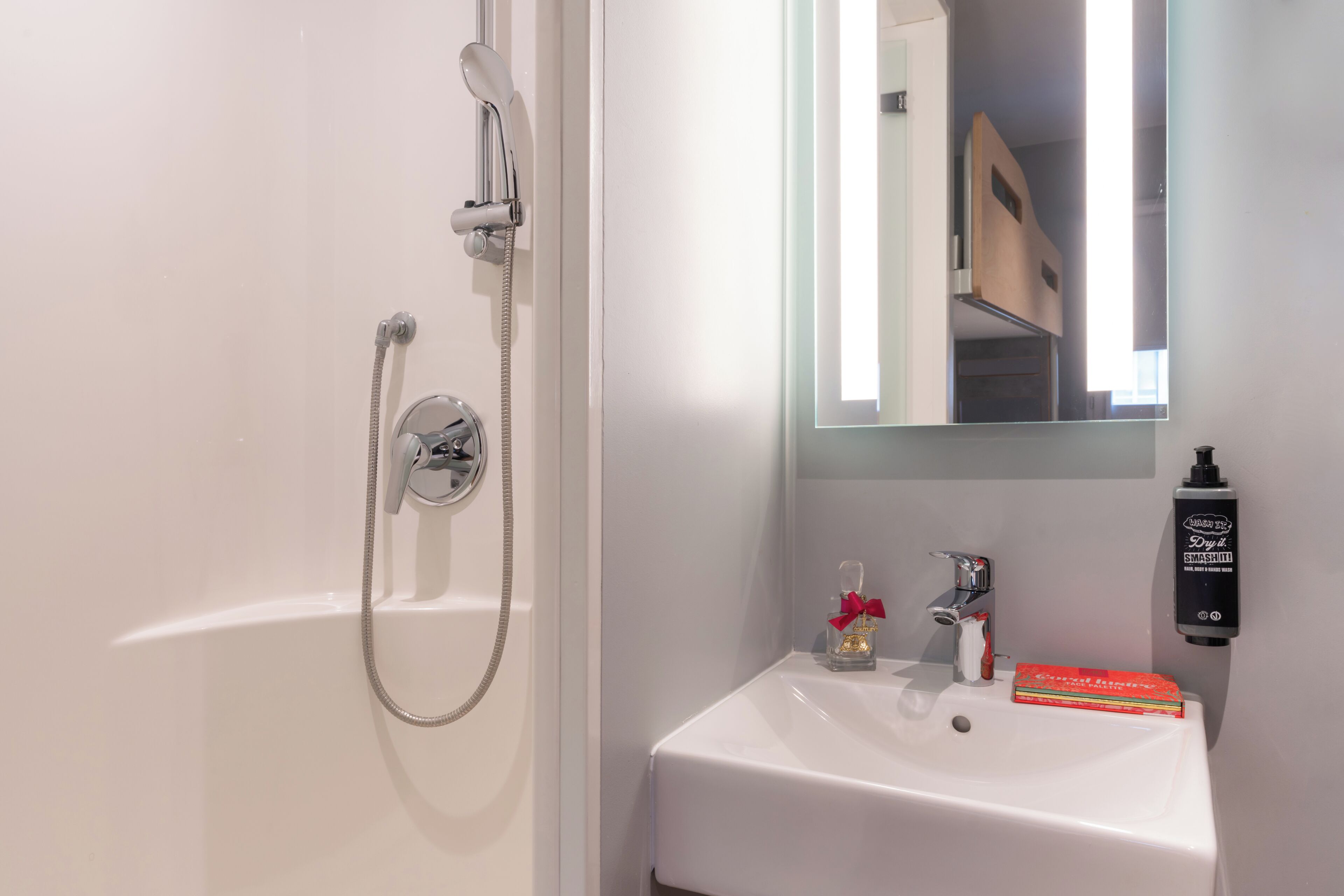 A bathroom with a white bathtub, a handheld showerhead, a sink with a mirror above it, and a bottle of hand soap on the wall.