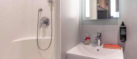 A bathroom with a white bathtub, a handheld showerhead, a sink with a mirror above it, and a bottle of hand soap on the wall.