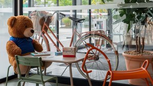 A teddy bear seated at a table with a book, a bicycle, and a rocking chair in a room with a tiled floor and a window. - ibis budget Marne la Vallée Val d'Europe (Montevrain)