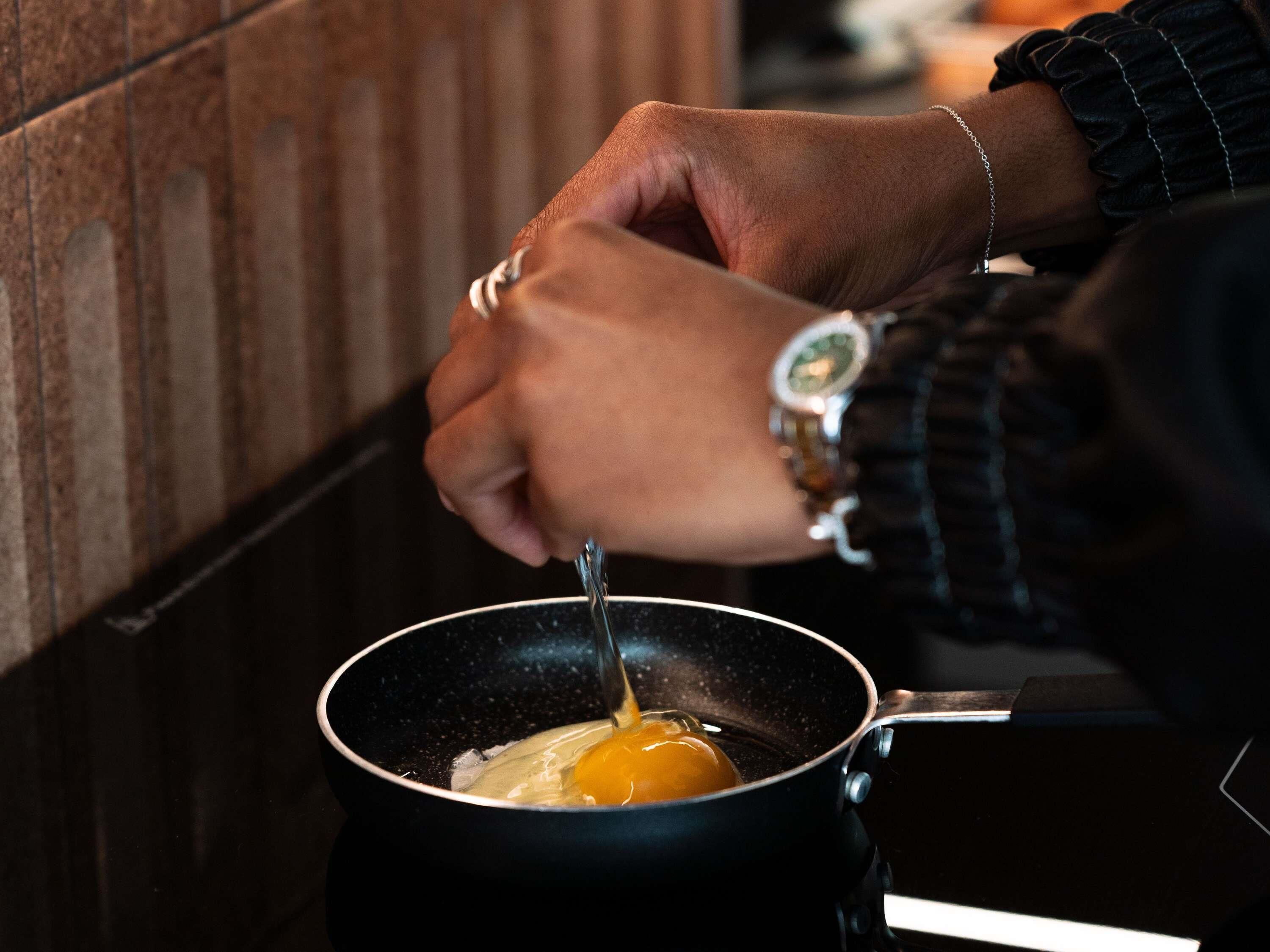 A hand cracking an egg into a black frying pan on a stovetop.