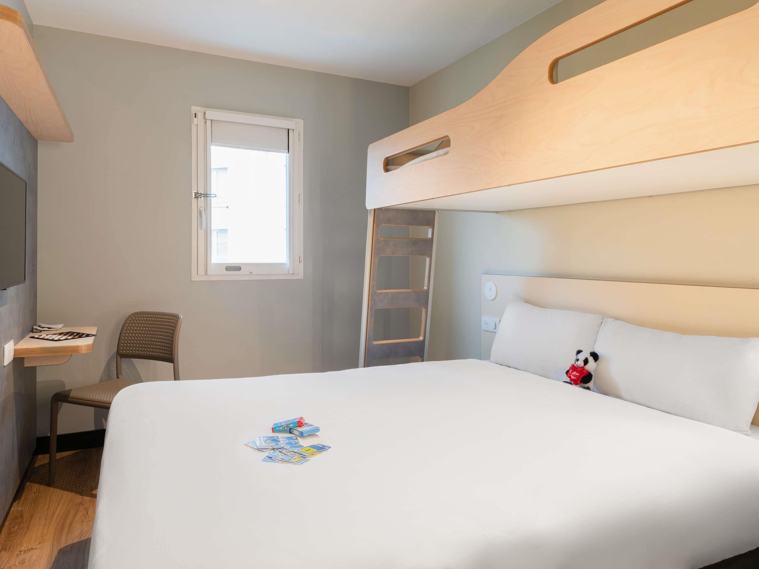 A hotel room with a bunk bed, a desk with a chair, a window, and a small table with a board game.