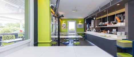 Un restaurant moderne avec des tables blanches et des chaises vertes, un comptoir où sont exposés des plats, et une vue sur la verdure à l’extérieur.