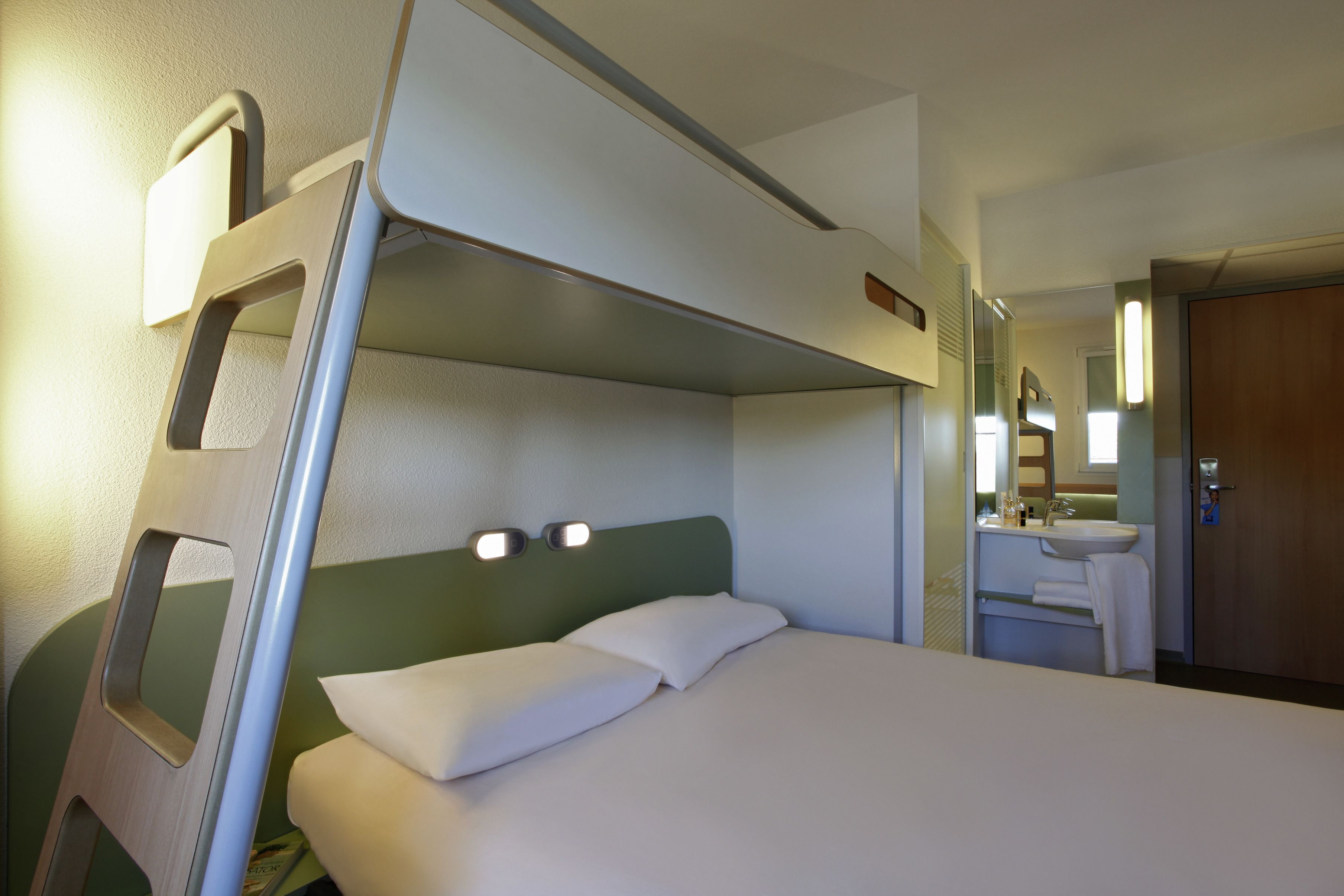 A single bed with a headboard, a bedside table, and a small mirror above the sink in a hotel room.