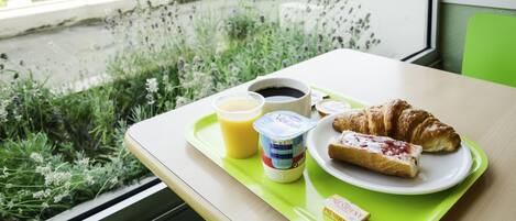 Bandeja de comida con croissant, jugo, yogur y café, colocada sobre una mesa junto a una ventana con vista a follaje y un sendero.