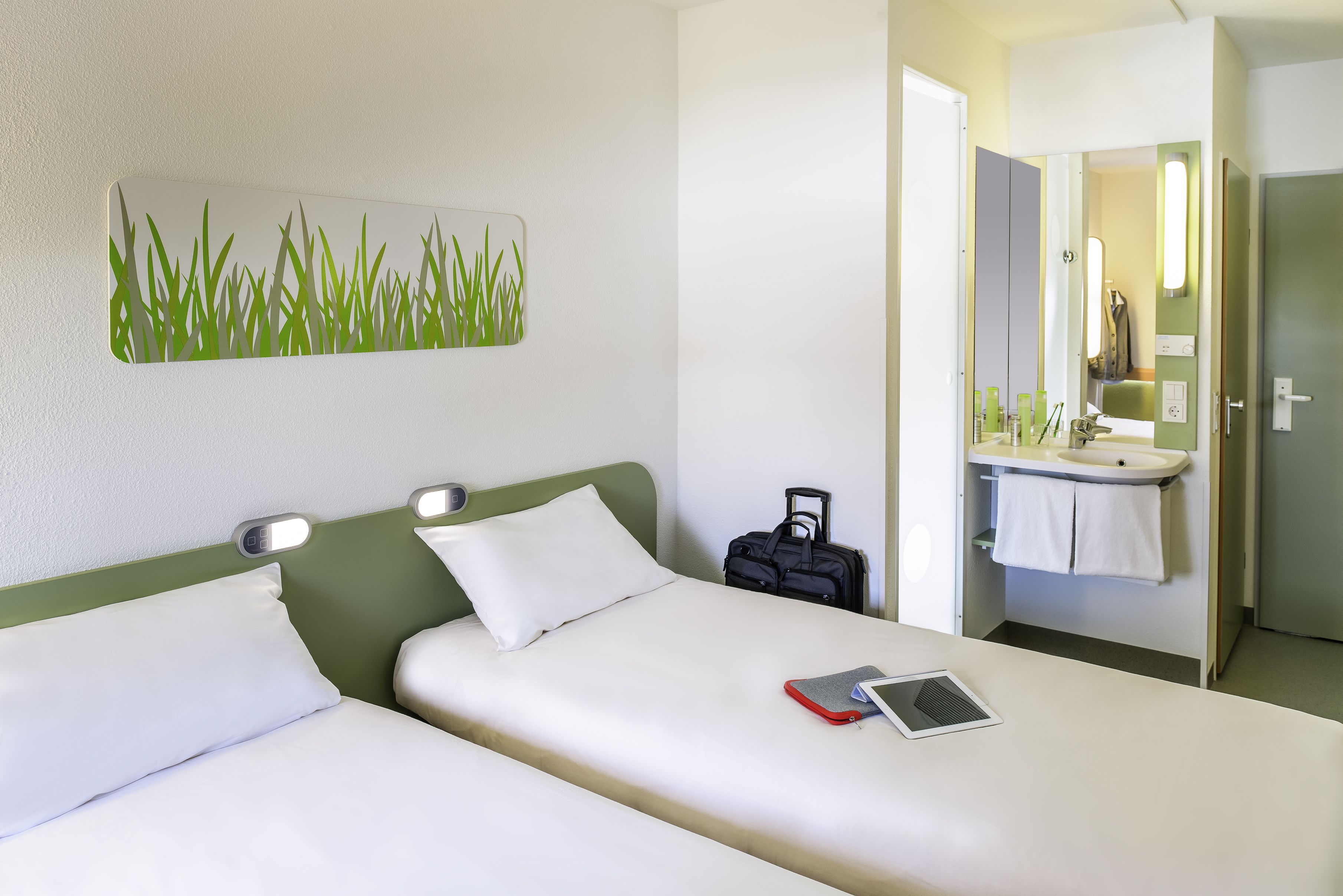 Une chambre d’hôtel avec deux lits, une table de chevet avec une lampe, un petit bureau avec une chaise, une salle de bain avec un lavabo et un miroir, et une œuvre d’art représentant de l’herbe, accrochée au mur.