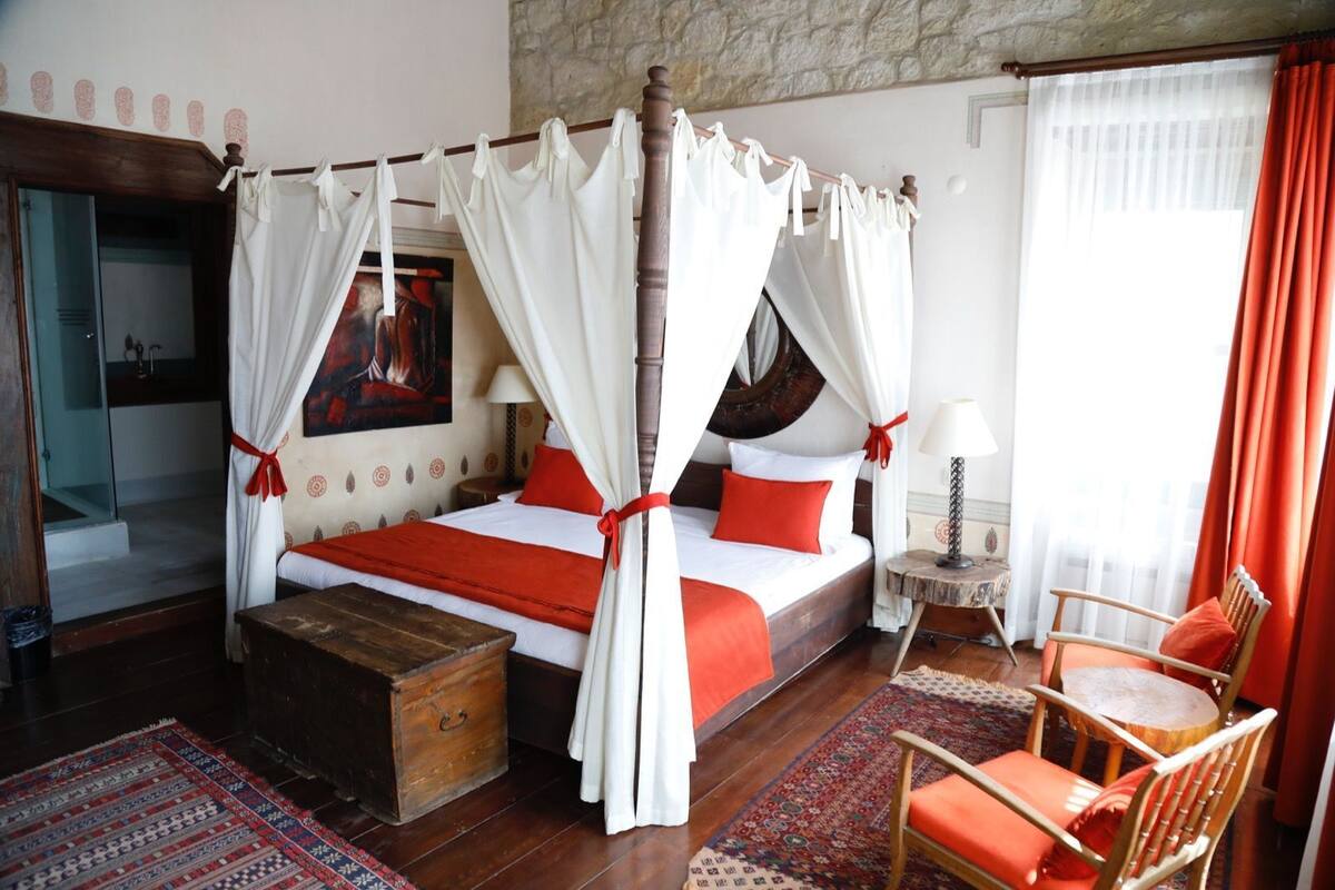 Una cama con dosel con ropa de cama blanca y roja, un cofre de madera, una mesita redonda con una lámpara, una silla y una alfombra estampada.