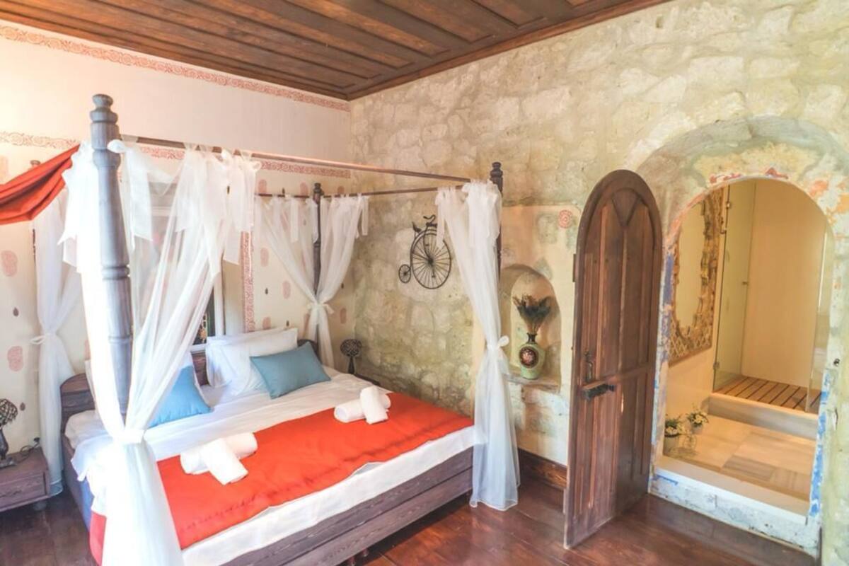 Una cama con dosel, ropa de cama blanca y una colcha roja, una pared de piedra y una puerta de madera.