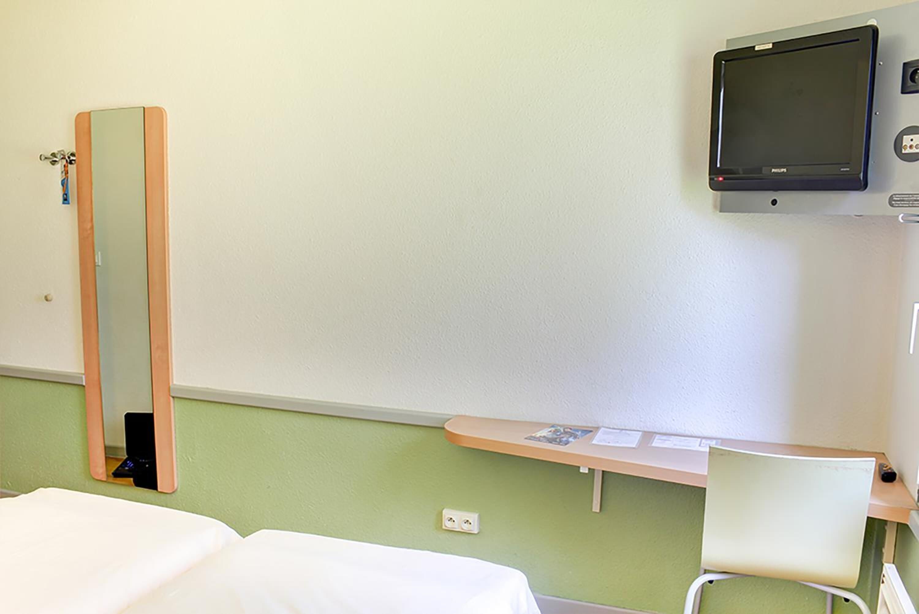 Ein Hotelzimmer mit Bett, Sessel, Schreibtisch, Spiegel und Wandfernseher.