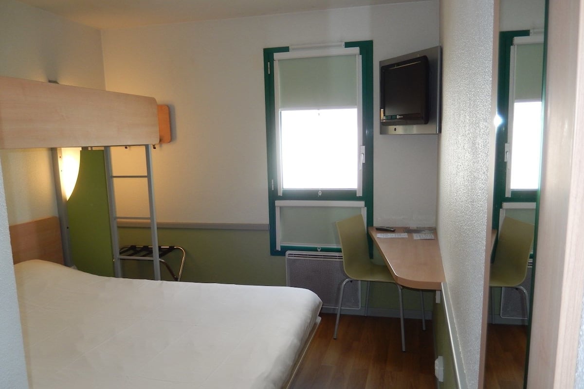 Une chambre d  ’hôtel compacte comprenant un lit superposé, un bureau avec une chaise, une télévision et une fenêtre.