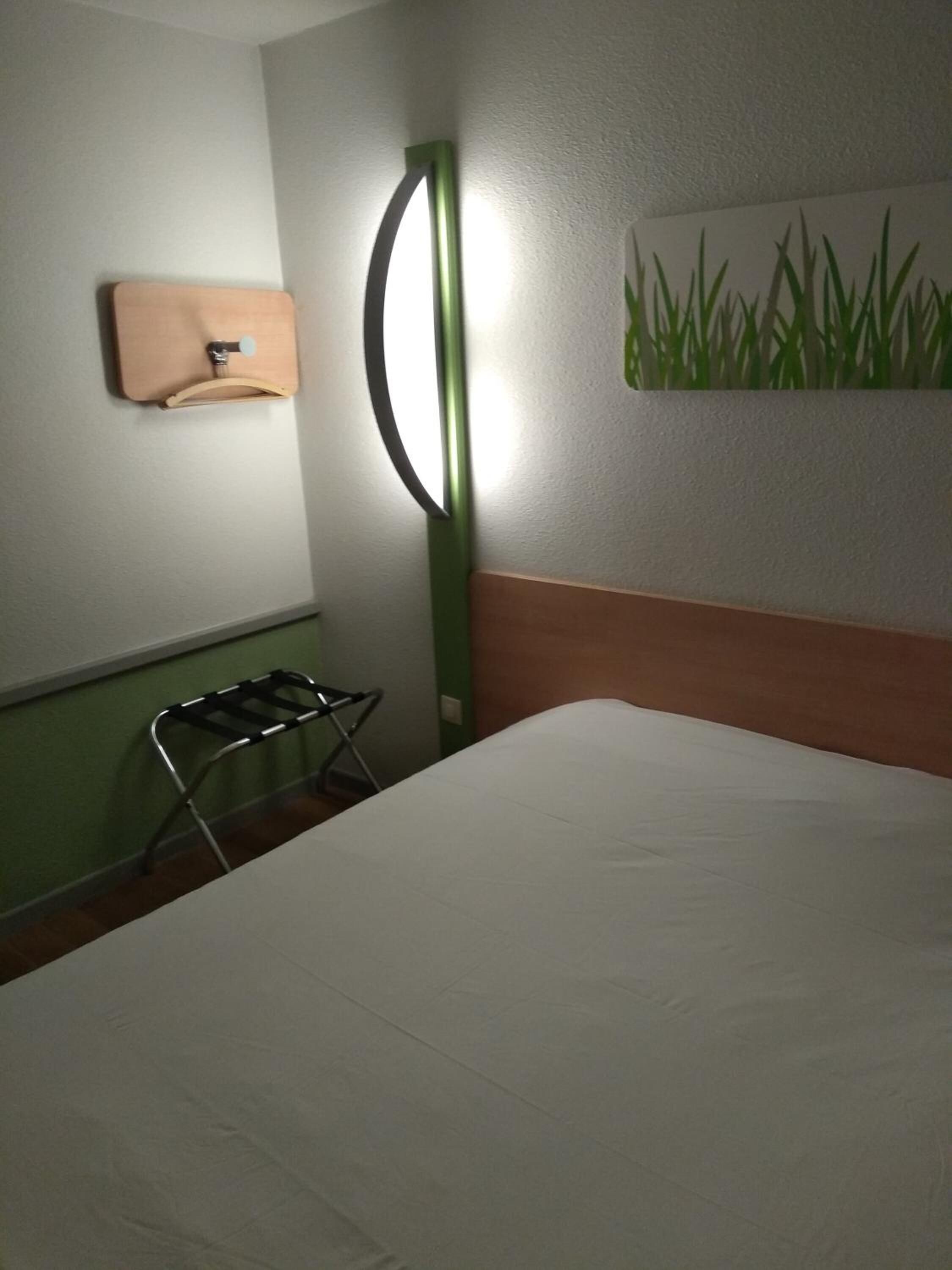 A hotel room with a bed, a wall-mounted towel rack, a green floor lamp, and a painting of grass on the wall.